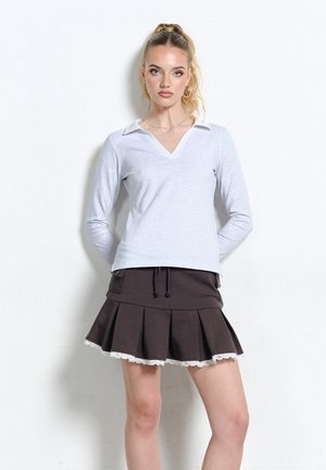 Młoda kobieta z blond kucykiem, ubrana w białą koszulę z długim rękawem i ciemną plisowaną spódnicę z koronkowym wykończeniem, stojąca na tle białej ściany.