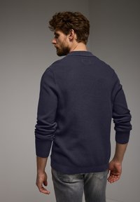 Mörkblå, texturerad stickad tröja med sjalkrage och långa ärmar, buren över ljusgrå jeans, vilket visar en avslappnad passform.