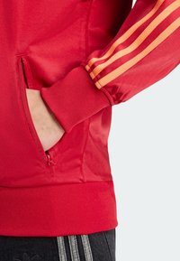 Röd jacka med dragkedja och oranga ränder på ärmarna. Tillverkad av ett slätt material, med ribbat mudd och sidoficka.