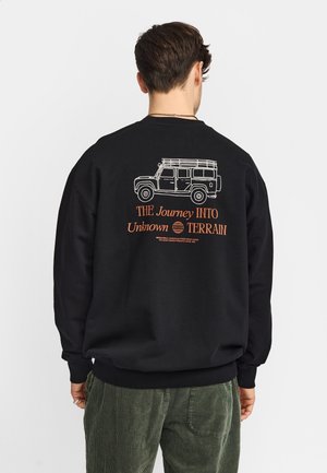 Man draagt een zwart sweatshirt met een witte schets van een offroadvoertuig en oranje tekst "DE Reis NAAR Onbekend TERREIN" op de rug, staand tegen een witte achtergrond.
