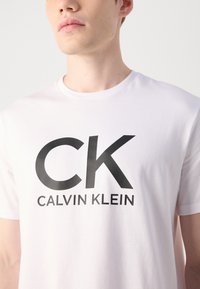 Witte katoen t-shirt met een groot zwart 'CK' en 'CALVIN KLEIN' logo gedrukt aan de voorkant. Klassieke ronde hals en korte mouwen.