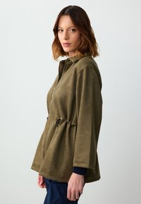 Giacca verde oliva con chiusura a zip, vita con cordino e morbido tessuto simile alla pelle scamosciata. Presenta tasche laterali e polsini a costine.