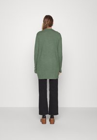 Lange groene cardigan van een zachte gebreide stof, met een geribbelde zoom, open voorkant en lange mouwen. Draagwijd over zwarte broek en hakken.