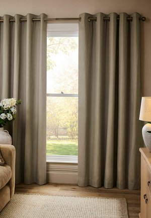 Beige Vorhänge teilweise geöffnet am Fenster, das den Garten draußen zeigt, neben einem beigen Sofa mit weißen Blumen und einer Lampe auf einem Holzschrank.