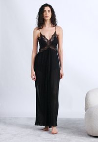 Γυναίκα με σγουρά μαλλιά στέκεται ξυπόλητη φορώντας ένα μακρύ μαύρο φόρεμα με δαντέλα και πιέτες, μπροστά σε ένα απλό λευκό φόντο.