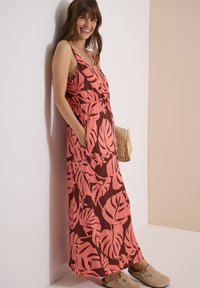 Femme souriante portant une robe longue sans manches avec un motif de feuilles roses et brunes, des sabots beige, tenant un sac à main tressé, appuyée contre un mur.