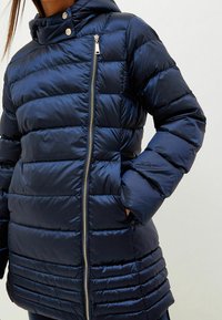 Námořnicky modrá péřová bunda s horizontálním pro�šíváním, zapínáním na zip, vysokým límcem a druky. Vyrobená z lesklého, lehkého materiálu.