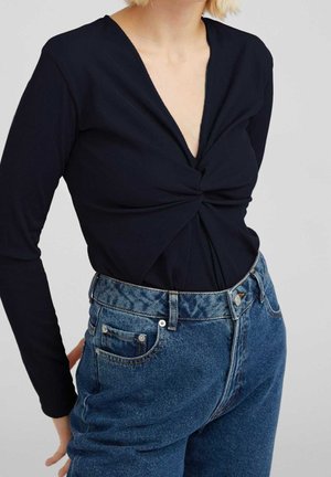 Kobieta nosząca czarny, długi rękaw z zakręconym przodem topu, włożony w wysokie niebieskie dżinsy, na jednolitym tle.