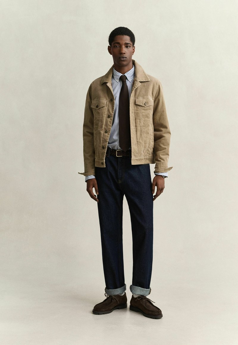 Junger Mann, der eine beige Jacke über einem gestreiften Hemd mit dunkler Krawatte, dunkle hochgekrempelte Jeans und dunkelbraune Schnürschuhe trägt und vor einem schlichten Hintergrund steht.