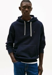 Marine hoodie met witte trekkoorden, kangoeroezak, geribbelde manchetten en een klein logo op de borst. Gecombineerd met blauwe jeans.