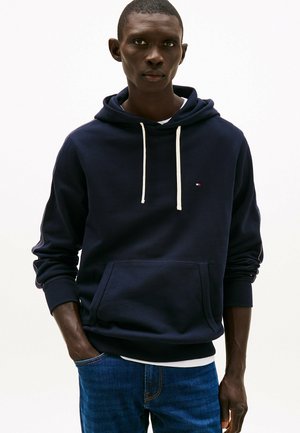 Marine hoodie met witte trekkoorden, kangoeroezak, geribbelde manchetten en een klein logo op de borst. Gecombineerd met blauwe jeans.