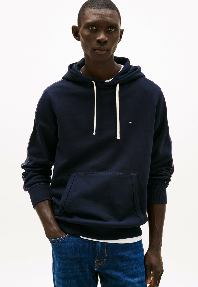 Marine hoodie met witte trekkoorden, kangoeroezak, geribbelde manchetten en een klein logo op de borst. Gecombineerd met blauwe jeans.