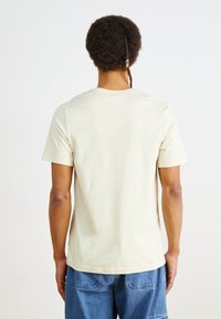 Tricou din bumbac light beige, cu guler rotund, mâneci scurte și o croială lejeră, expus din spate. Textură netedă, fără grafică.