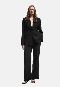 Sort skræddersyet jakkesæt med en enkelt-knap blazer med notch revers og brede bukser, suppleret med strappy sorte hæle.