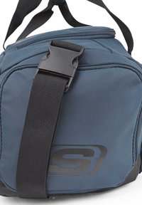 Bolsa deportiva azul hecha de material duradero, con una correa ajustable negra gruesa y un cierre de plástico negro. Presenta un logotipo discreto.