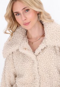 Cappotto bouclé beige con una texture fluffy, colletto ampio e chiusura a un bottone. Indossato sopra un top chiaro, con delicati accenti di gioielli.