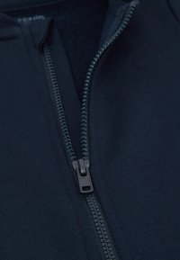 Navy zippered jacket featuring a textured fabric, smooth zipper, and ribbed collar. Close-up highlights zipper detail and material texture.