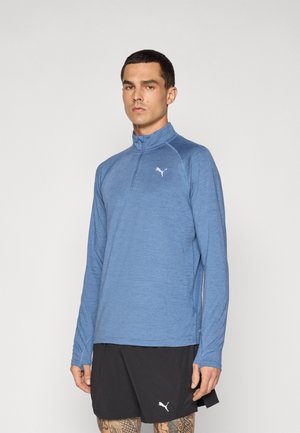 Light blue long-sleeve technical shirt with a quarter zip, featuring a white logo on the chest and raglan sleeves, paired with black shorts.