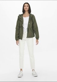 Giacca a vento verde oliva con cappuccio, bottoni frontali, polsini elasticizzati e una vestibilità rilassata, indossata sopra una maglietta bianca e abbinata a pantaloni bianchi e sneaker.