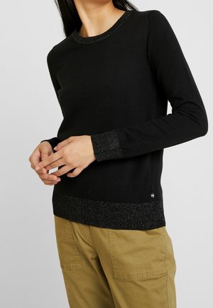 Femme portant un pull noir à manches longues avec poignets et ourlet pailletés, associé à un pantalon marron clair sur un fond uni.