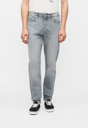 Homme portant un jean droit gris clair, un t-shirt blanc à motif graphique et des baskets basses noires avec des lacets blancs sur un fond uni.
