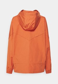 Orange huva jacka med en texturerad tyg, med rak skärning och elastiska ärmslut, tydligt synliga sömdetaljer på ryggen.