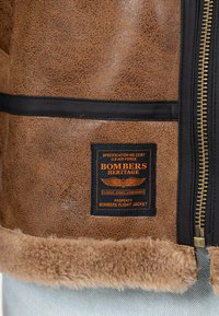 Braune Kunstleder-Flugjacke mit Schafsfellkragen, schwarzen Akzenten und einem Etikett mit orangefarbigem Text und einem Flügelgrafik.