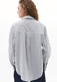 Donna con capelli ricci che indossa una camicia bianca a maniche lunghe con strisce nere verticali e orizzontali, abbinata a pantaloni scuri, che guarda a sinistra.