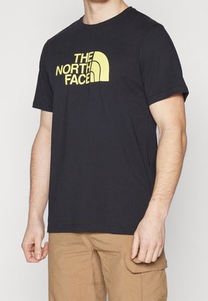 Homme portant un t-shirt noir The North Face avec un logo jaune et un short cargo beige, visible du cou jusqu'à mi-cuisse sur fond uni.