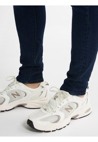 Bílé síťované běžecké boty s béžovými akcenty, s tkaničkami a černými podrážkami. Nosené s tmavě modrými skinny džíny, odkryté kotníky.