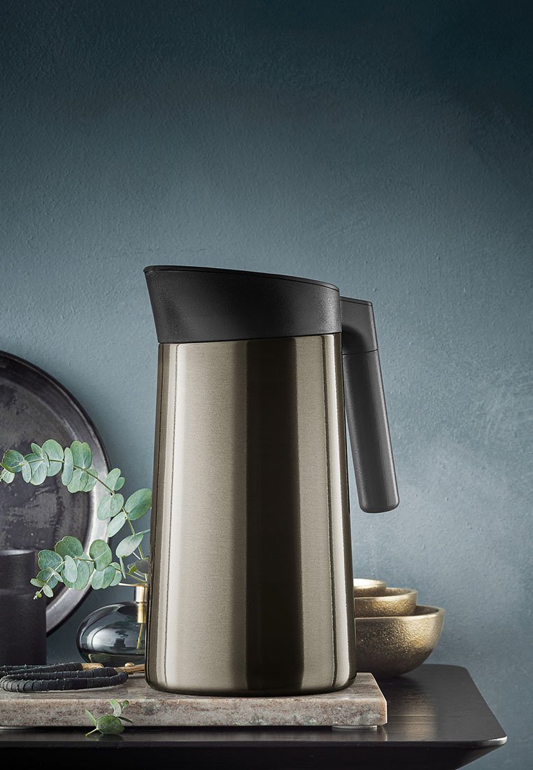 Kaffeekanne aus Edelstahl mit gebürsteter Oberfläche, schwarzem Deckel und Griff sowie tapered Design. Umgeben von Grünpflanzen und Dekorationsobjekten.