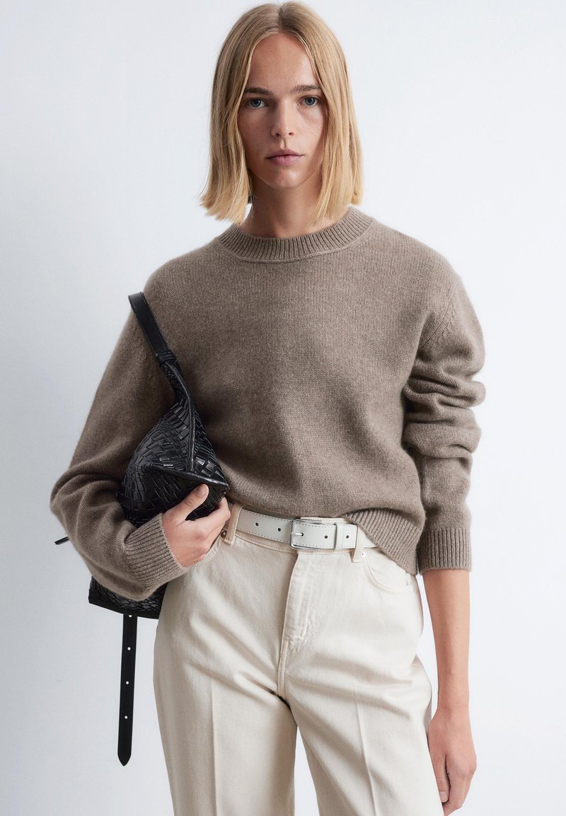 Frau mit glattem, blonden Haar, die einen taupe-farbenen Strickpullover, cremefarbene High-Waist-Hosen mit weißem Gürtel trägt und eine schwarze geflochtene Ledertasche hält.