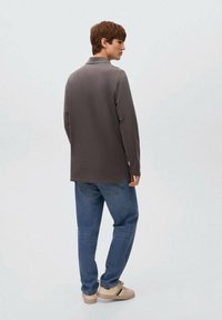 Junger Mann steht seitwärts, trägt ein dunkelgraues Langarmshirt, blaue Jeans und beige Sneaker mit grünen Streifen.