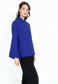 Blå blousonskjorta med långa trumpetärmar, knapprad fram och sidslitsar. Tillverkad av lätt tyg med en mjuk yta.