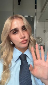 Osoba o blond włosach, ubrana w jasnoniebieską koszulę i ciemny krawat w wzory, pokazująca dłoń z czarnym lakierem do paznokci, na tle płytek.