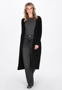 Donna che indossa un lungo cardigan nero sopra una maglia grigia, jeans neri a vita alta e scarpe nere, in piedi contro uno sfondo bianco.