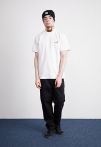 White short-sleeve t-shirt with a small orange and yellow logo, paired with black pants and black sneakers, worn with a black beanie hat.