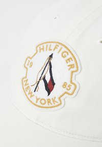 Casquette blanche avec un badge brodé. Le badge affiche "HILFIGER," "NEW YORK," et "1985," avec un design comprenant des accents rouges, noirs et jaunes.