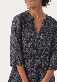 Marineblauwe bloemen-tunic met 3/4 mouwen, met een geplooide halslijn. Het ontwerp bevat lichtgekleurde bloemmotieven op een donkere achtergrond.