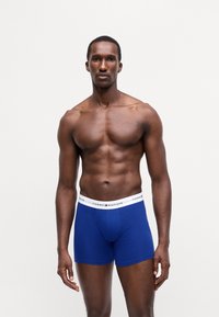 Modri bombažni bokserice z belim pasom, na katerem je napis "TOMMY HILFIGER". Model ima atletsko postavo in je brez majice.