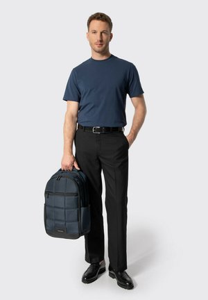 Hombre vistiendo una camiseta azul marino y pantalones negros, sosteniendo una mochila acolchada azul marino con una mano, de pie frente a un fondo liso.