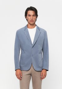 Man wearing light blue blazer over white shirt and beige pants, standing against plain white background.