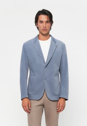 Man wearing light blue blazer over white shirt and beige pants, standing against plain white background.