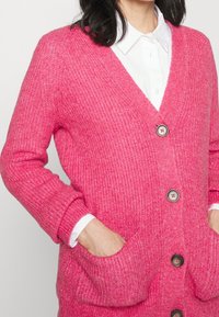 Rosa stickad cardigan med V-ringning, långa ärmar, två framfickor och kontrasterande knappar. Texturen är mjuk och något tjock.