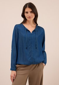 Femme aux longs cheveux noirs portant un blouse bleue à manches longues avec des cordons et un pantalon beige sur fond uni.