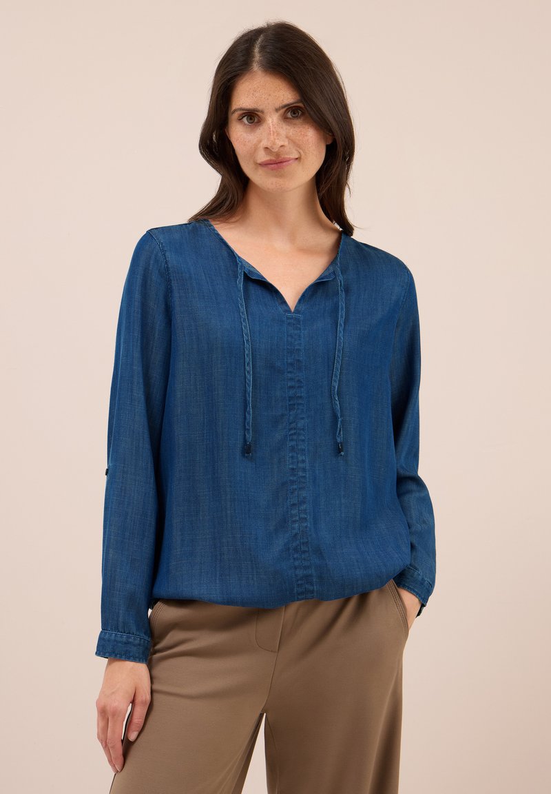 Femme aux longs cheveux noirs portant un blouse bleue à manches longues avec des cordons et un pantalon beige sur fond uni.