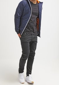 Giacca imbottita blu navy con zip bianca e interno arancione, t-shirt nera, jeans scuri e scarpe da ginnastica bianche. Abbigliamento casual.