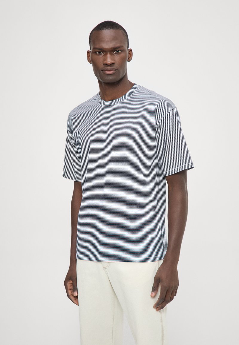 Camiseta a rayas en azul y blanco, de manga corta, cuello redondo, corte relajado, hecha de algodón. El modelo la lleva con pantalones en tonos claros.