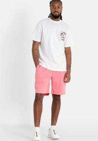 Wit katoenen T-shirt met opdruk op de linkerborst, gecombineerd met roze korte broek. Model draagt witte sneakers met accenten. Eenvoudig ontwerp en casual pasvorm.