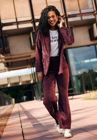 Completo in cotone a coste di un profondo bordeaux, composto da un blazer e pantaloni ampi, abbinato a una maglietta bianca e a delle sneakers bianche. Tessuto testurizzato visibile.